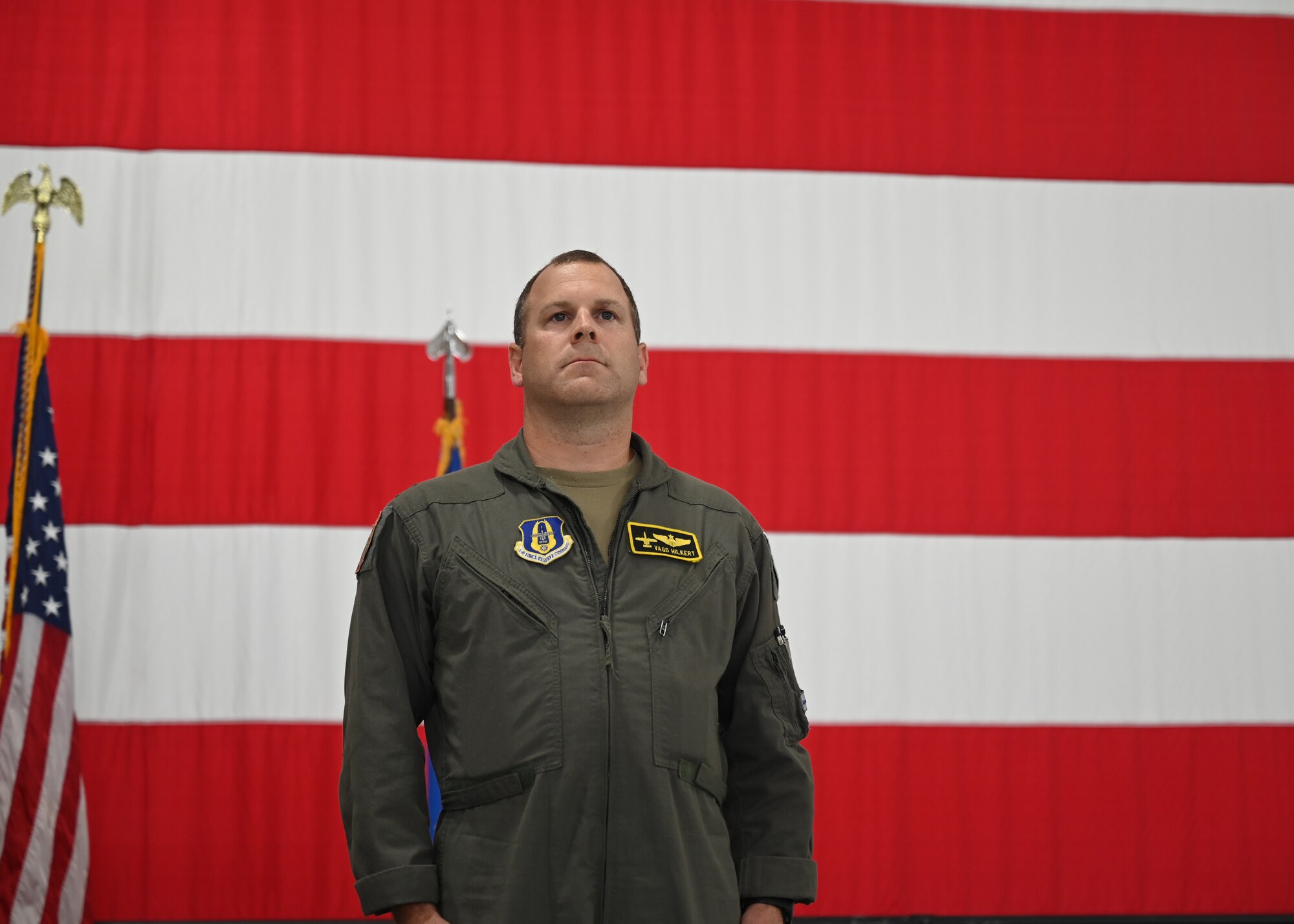 A man stands in front of a large American flag wearing green coveralls. The patch on his left breast reads "Vago Hilkert".