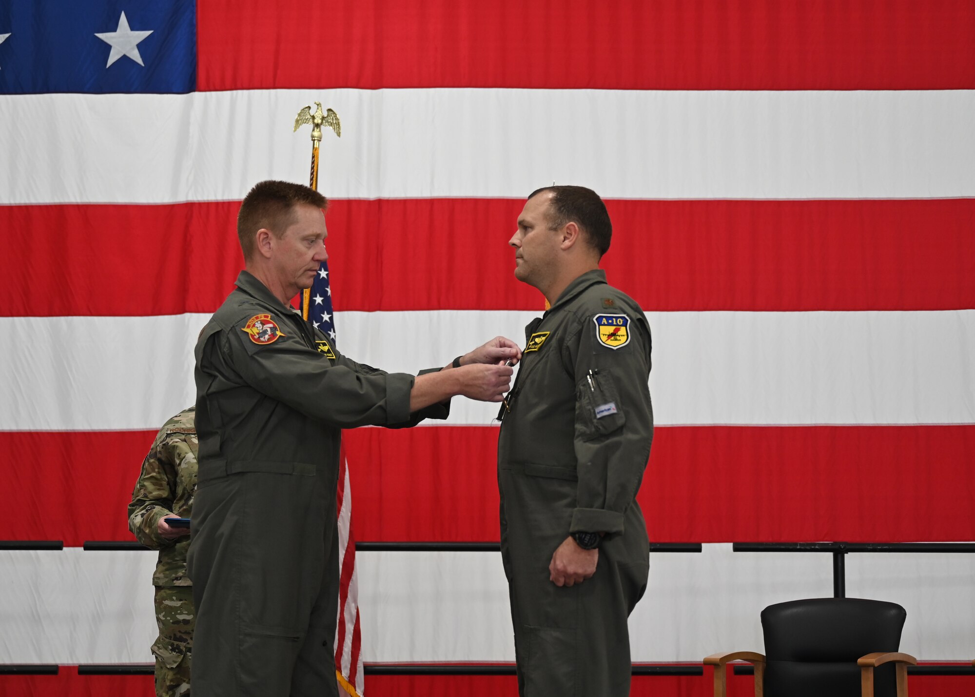 A man pins a medal to the chest of another man in front of a large American flag.