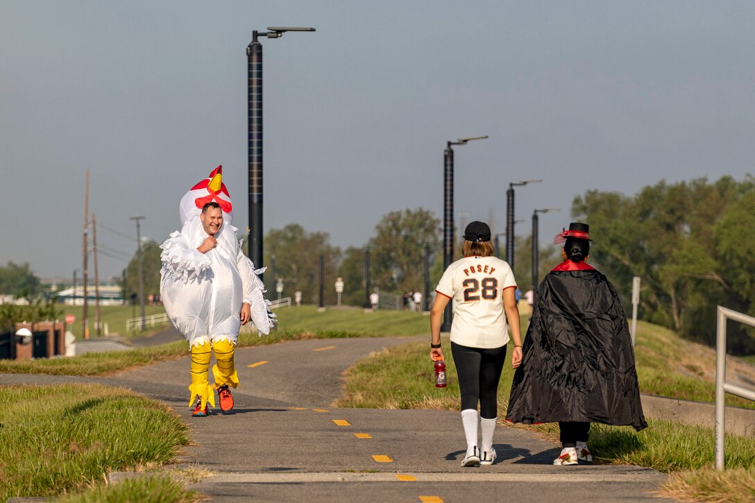 A person in a chicken costume walks on an asphalt patch toward two other people in costume.