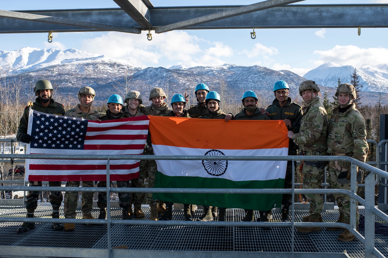 A group of soldiers pose for a photo holding two flags with mountains in the background.