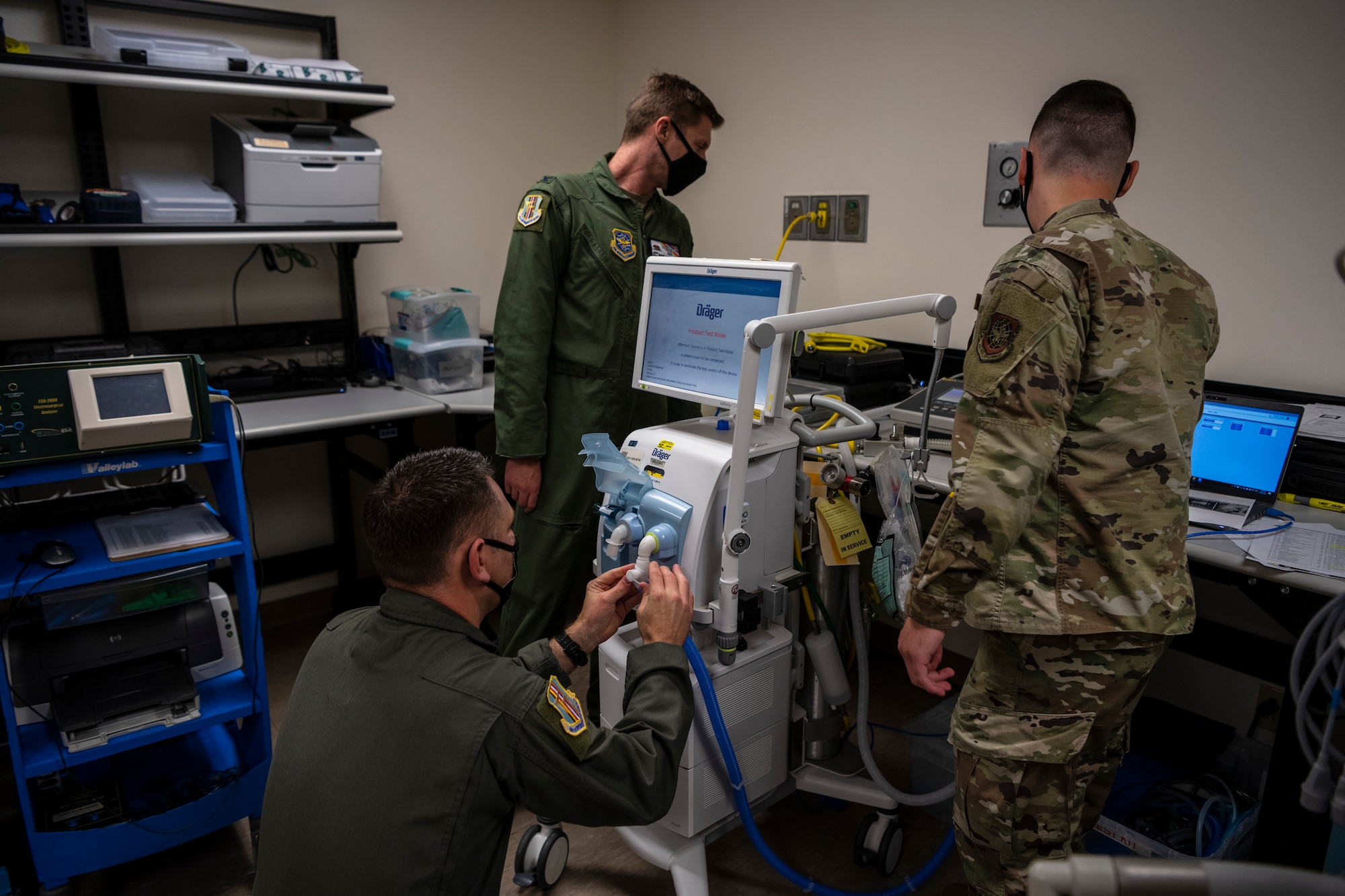 Men in uniform are crowded around a ventilator machine connecting parts to each other.