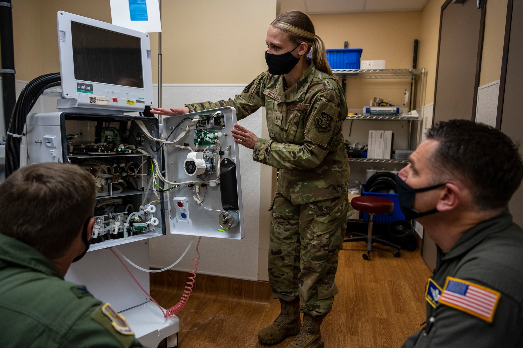 A woman in uniform shows a large white piece of medical equipment to two men in uniform.