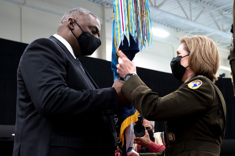 A man in a business suit passes a flag to a woman in military dress.