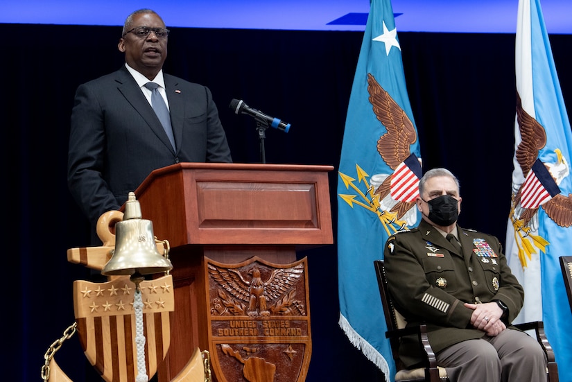 A man stands at a lectern while another man in a military uniform is seated to his left. A flag is in the background.