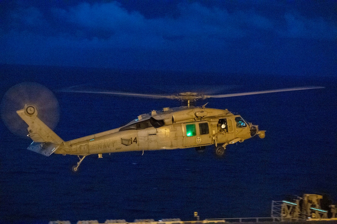 A helicopter prepares to land on an aircraft carrier at night.