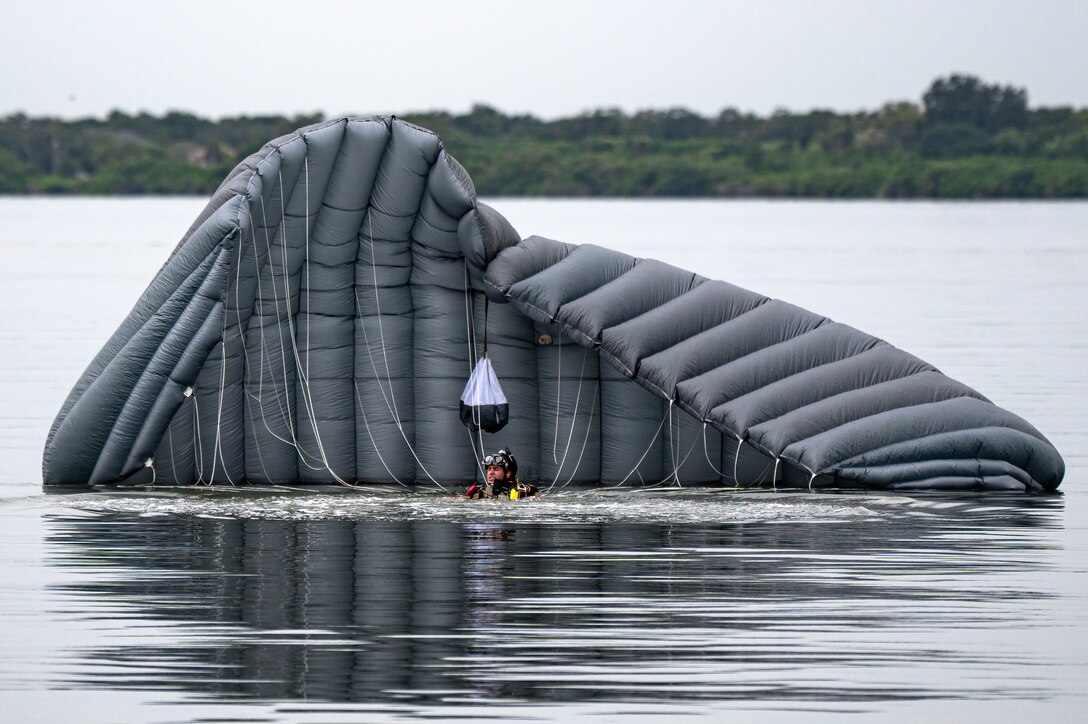 An airman stands in the water in front of a large deflated parachute.