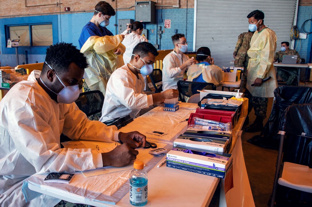 Sailors wearing face masks and gloves sit at a table preparing COVID-19 tests.