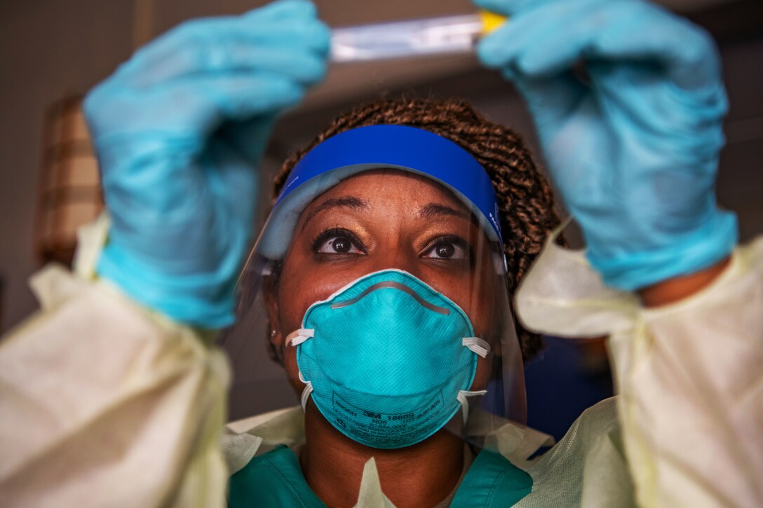 An airman wearing a face mask, gloves and medical gown holds up a test swab to check the label.