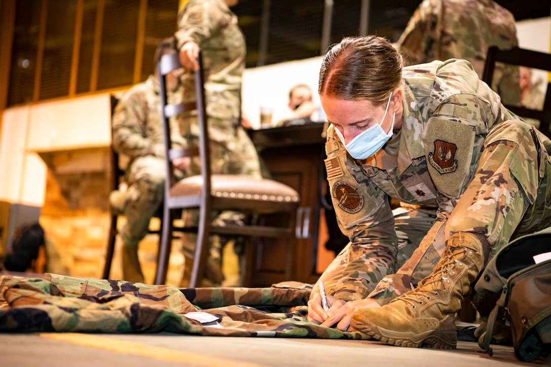 An airman writes on gear while sitting on the floor as fellow service members sit at a table in the background.