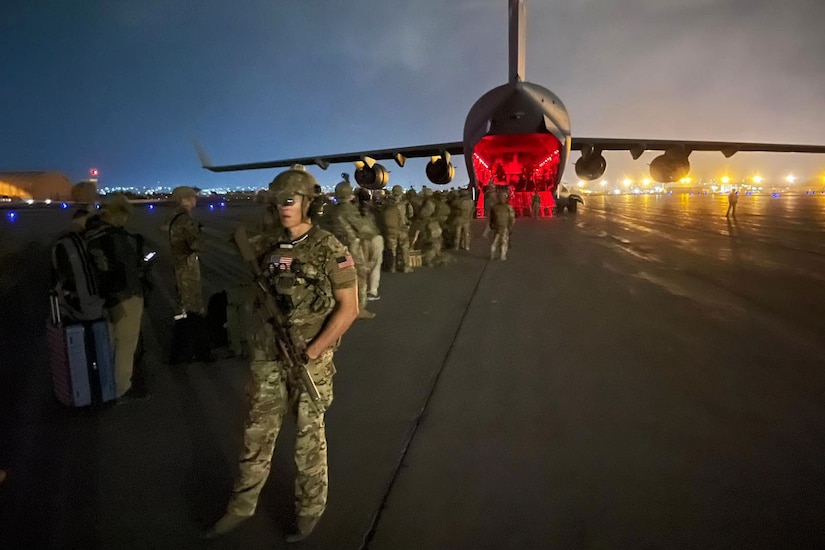 A soldier stands with a weapon a flightline as others stand in line by an open aircraft.