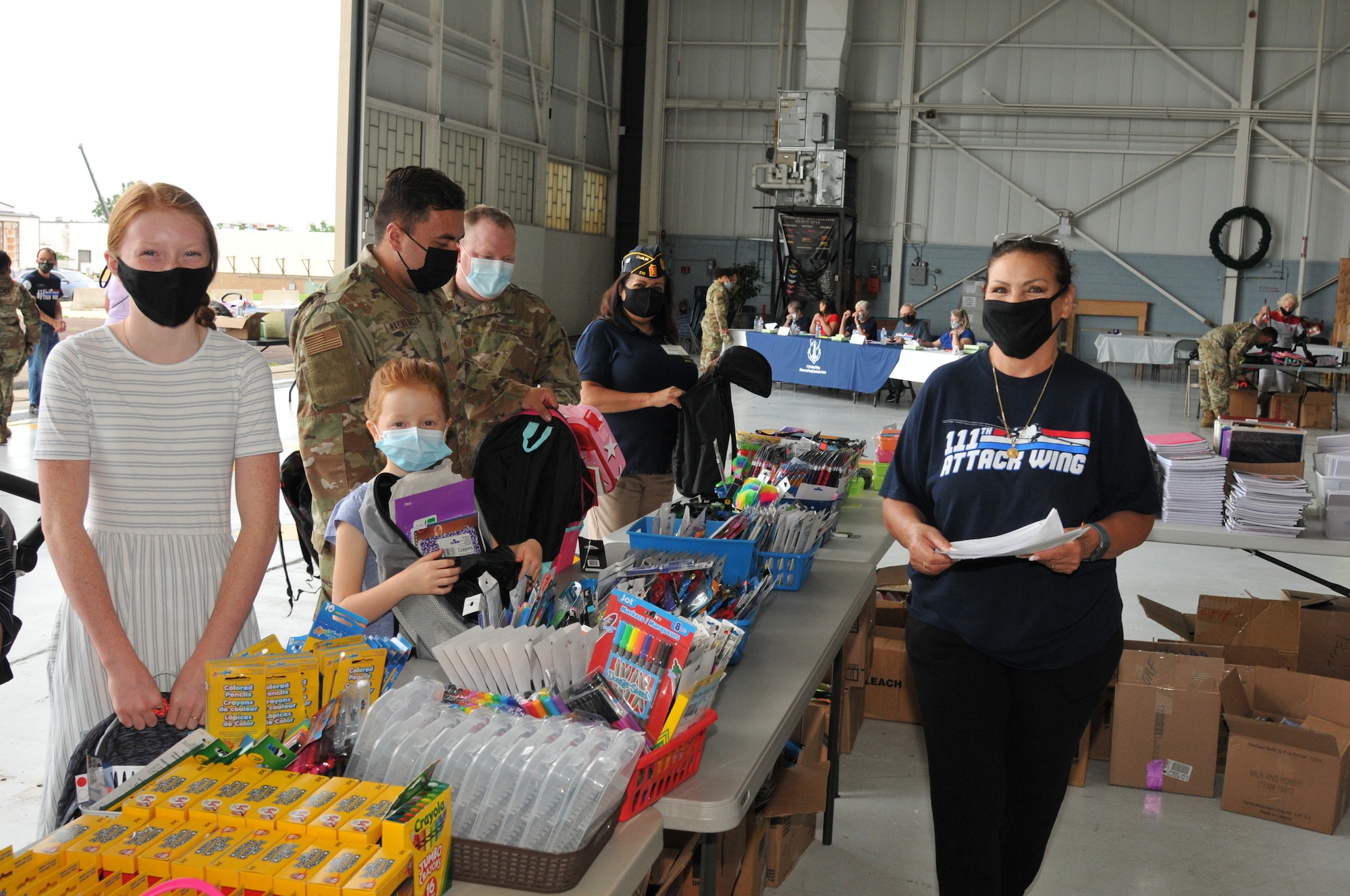 A woman in a mask poses next to a table of school supplies opposite a military family.