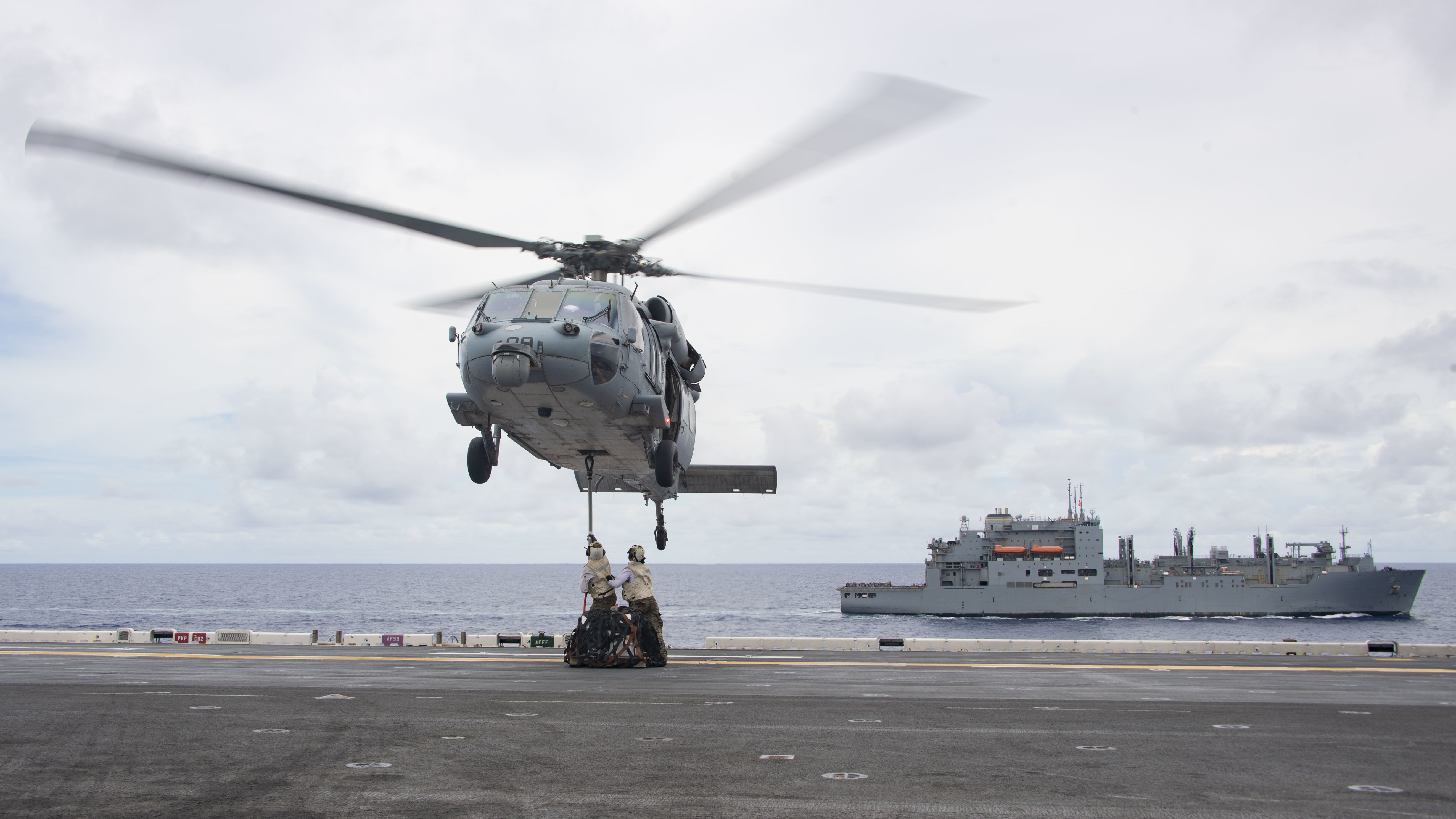 An MH-60S Sea Hawk conducts a vertical replenishment (VERTREP) with dry cargo and ammunition ship USNS Matthew Perry (T-AKE 9), back, and amphibious assault ship USS Essex (LHD 2).