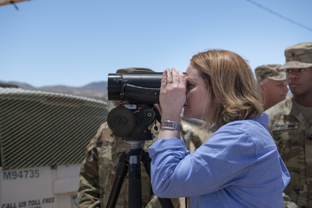 A woman uses a device to view something in the distance. A service member stands next to her.