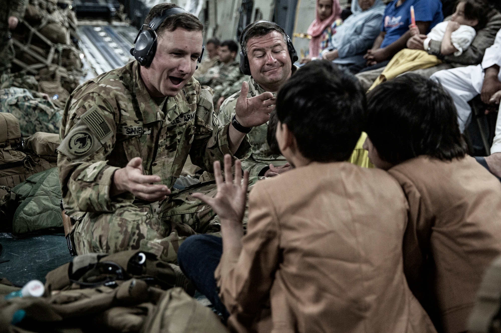 A soldier smiles and gesticulates while sitting on an aircraft floor with two youths.