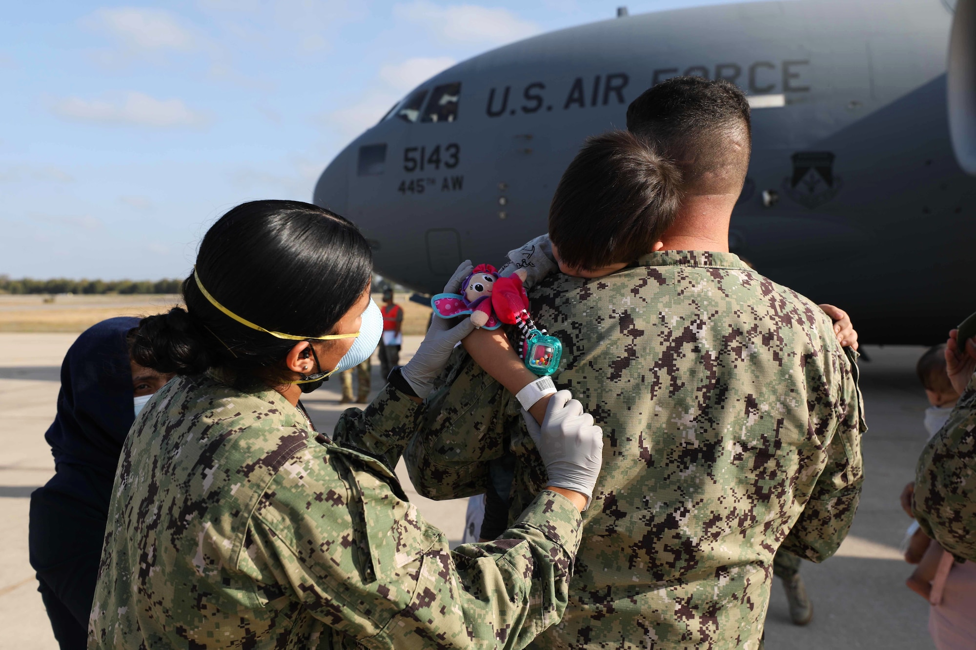 A sailor holds a young child as another hands the child a toy.