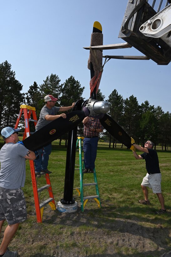 Four men help position a U.S. Air Force B-25 propeller display into place as it is hoisted up using a forklift on the grassy grounds of the North Dakota Veterans Home, Lisbon, N.D., Sept. 1, 2021.