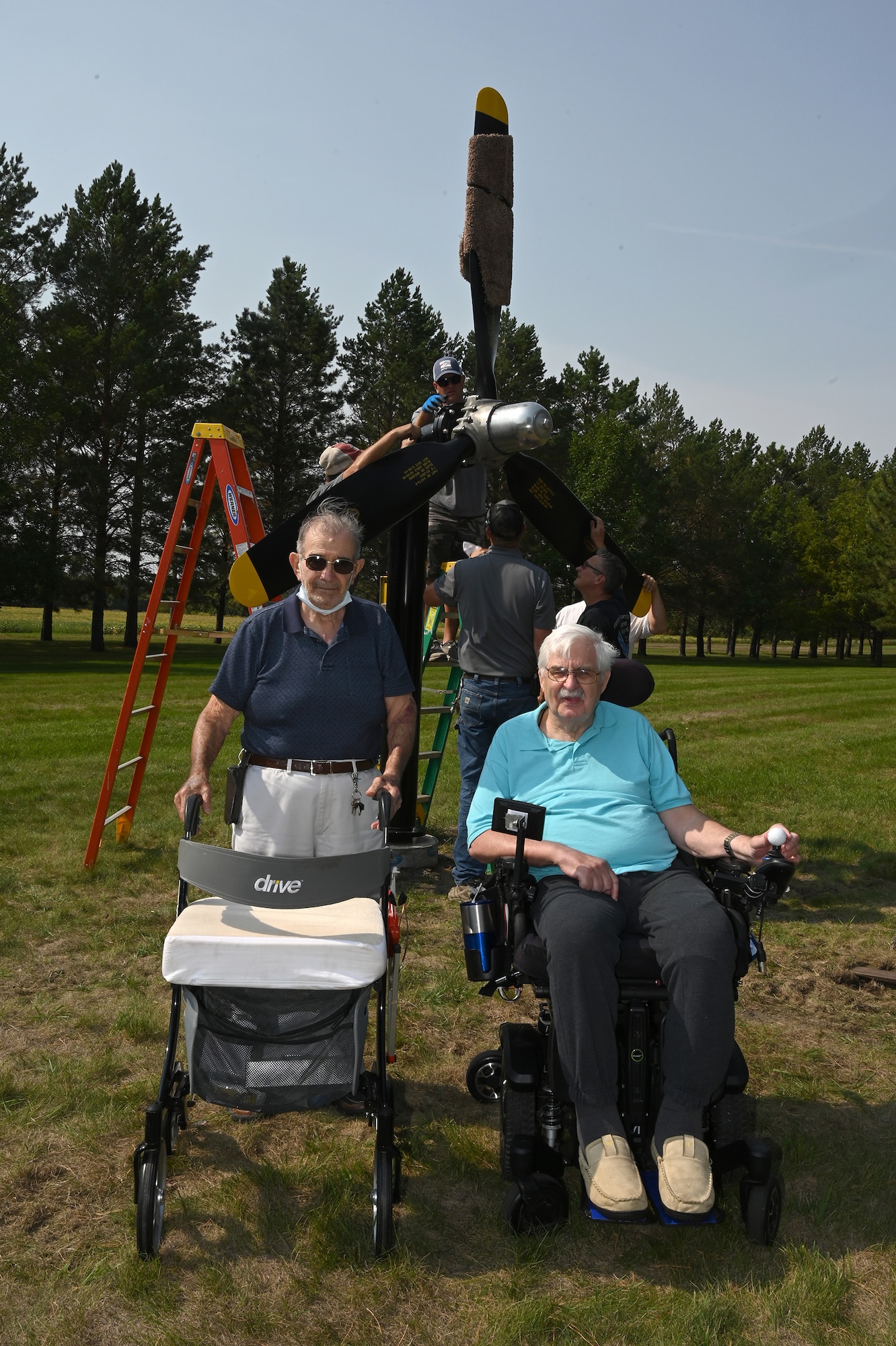 Two elderly men pose for a photo in front a U.S. Air Force B-25 propeller display being installed by four men on the grassy grounds of the North Dakota Veterans Home, Lisbon, N.D., Sept. 1, 2021.