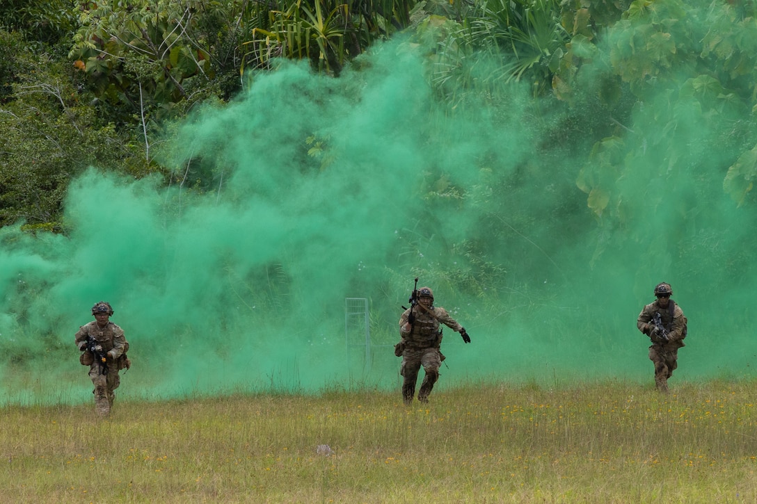 Three soldiers run across a field with green smoke behind them.