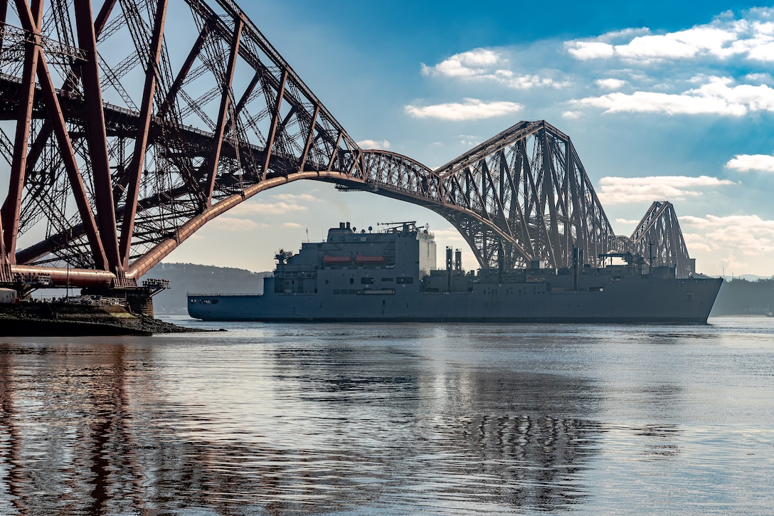 A military ships steams under a large bridge.