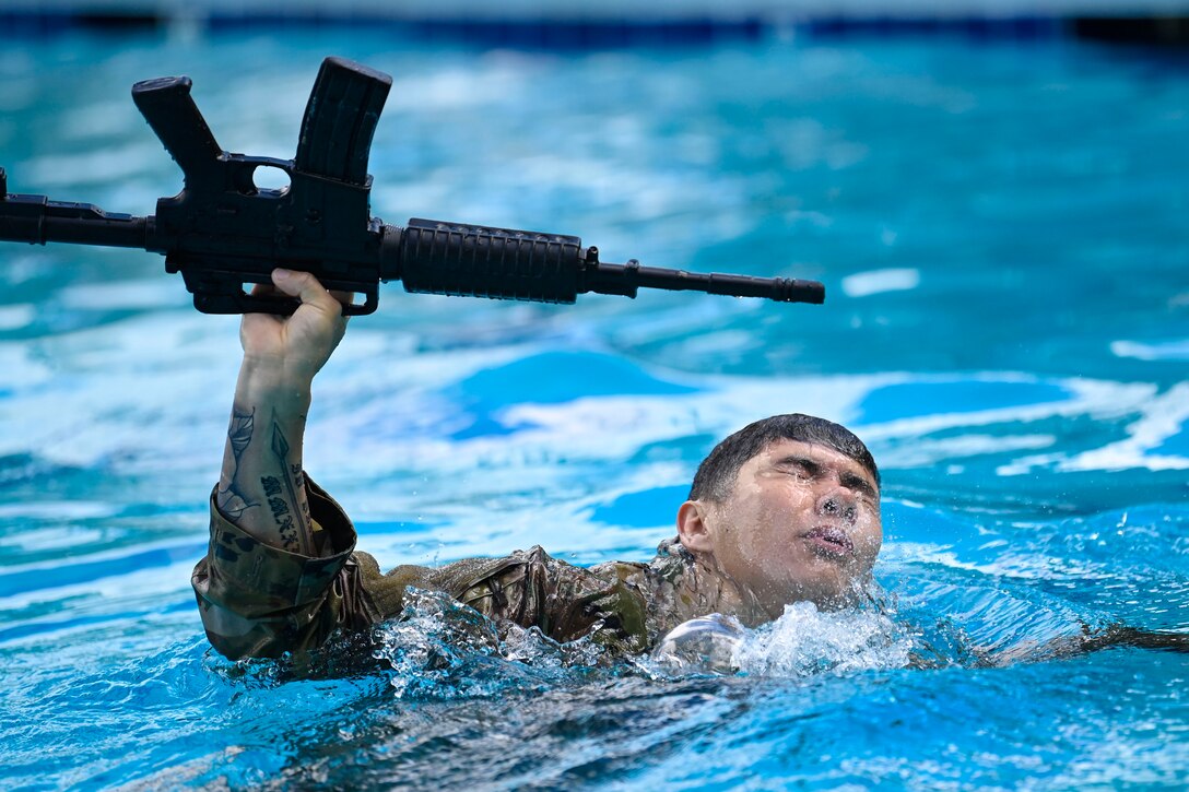 A soldier swims in a pool while carrying a rifle.