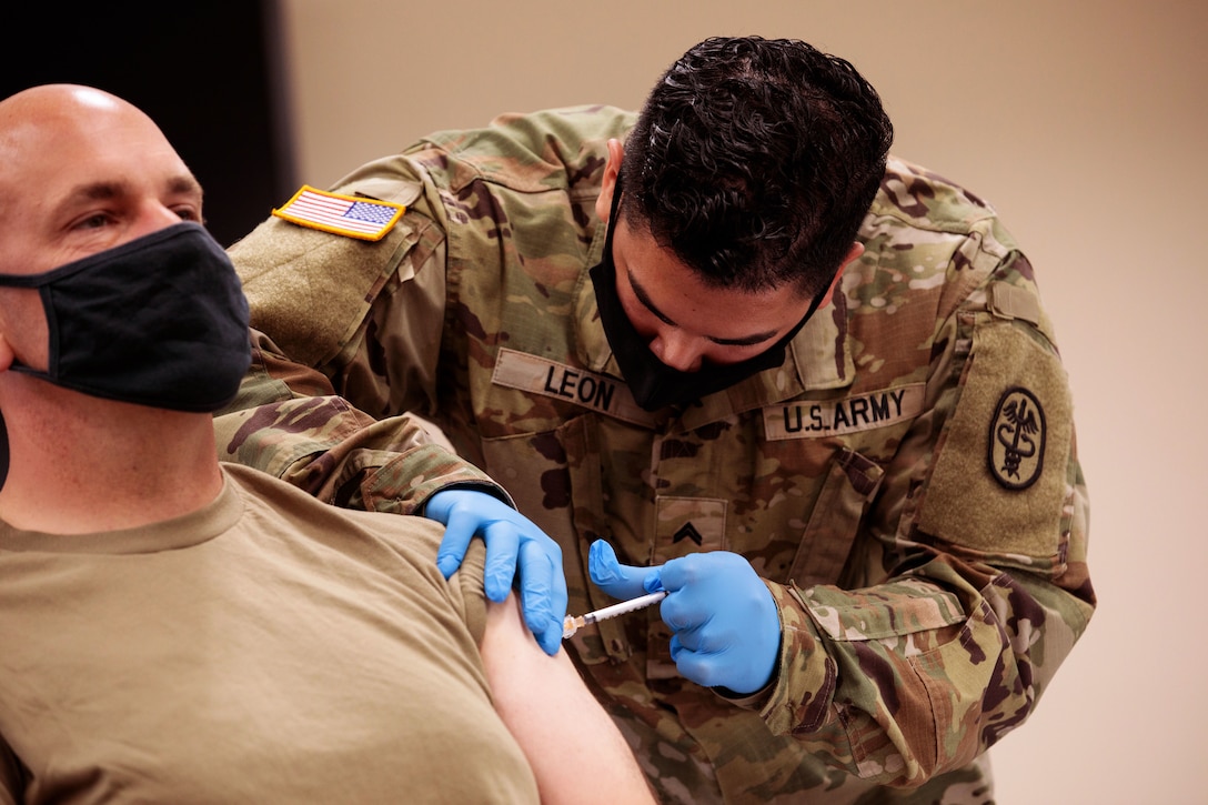 A soldier wearing a face mask and gloves holds a syringe while bending over to give another soldier wearing a face mask a vaccine.