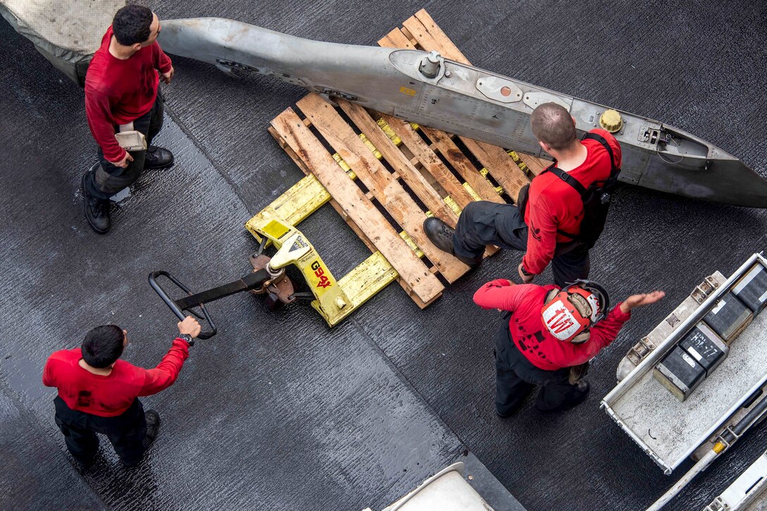 Four sailors in red, viewed from above, position a large metal object on a dolly.