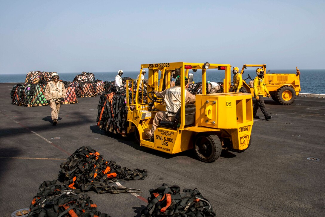 A service member steers a yellow forklift on a flight deck where others work.
