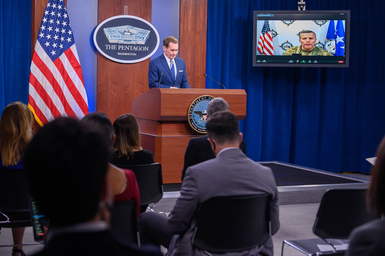 person on screen and person at lectern in briefing room.