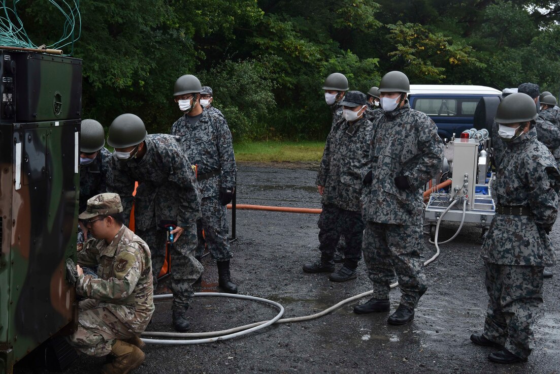 A U.S. military member crouches in front of a generator while Japanese military members watch.