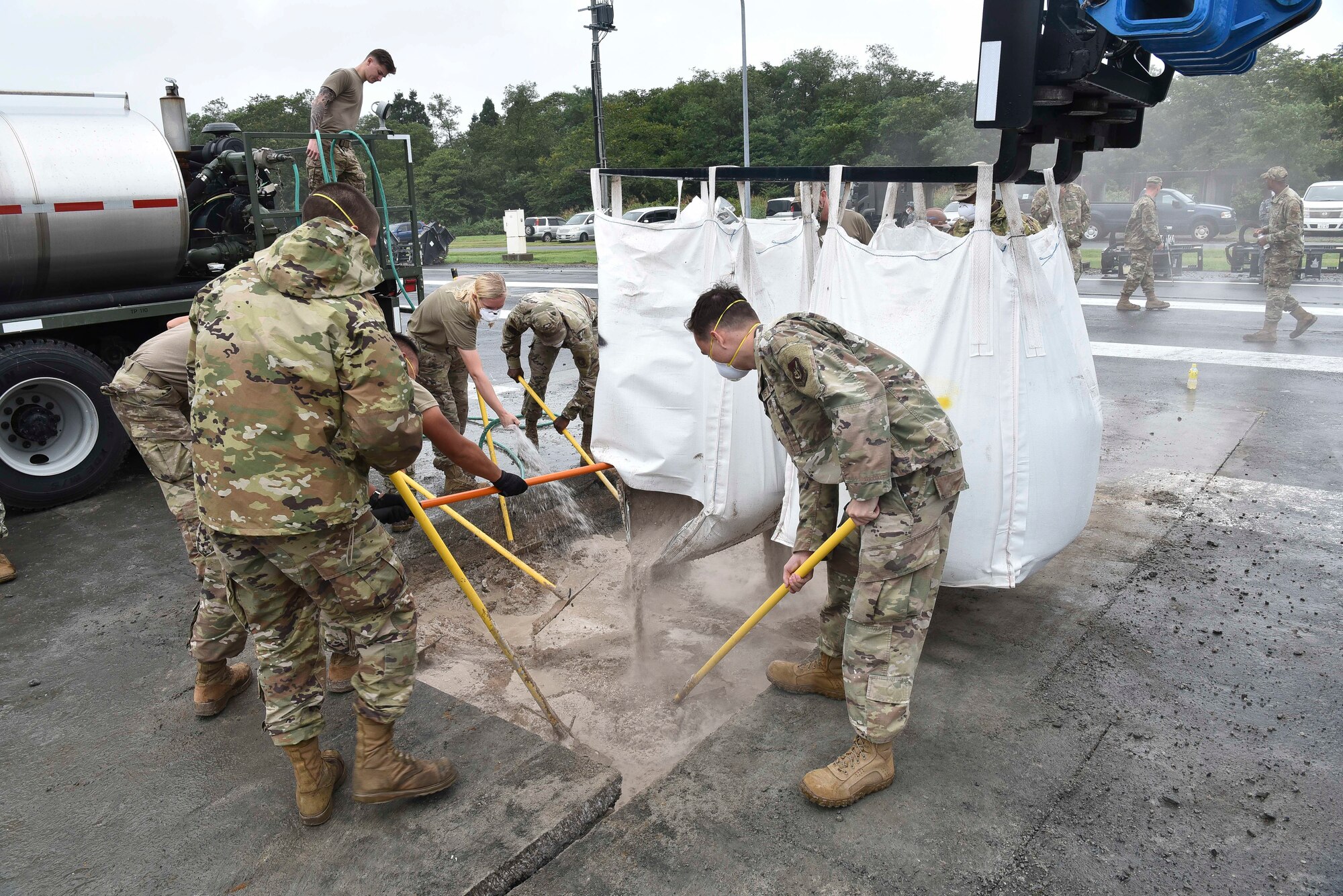 Military members scrape cement into a hole, from a bag held by a crane.