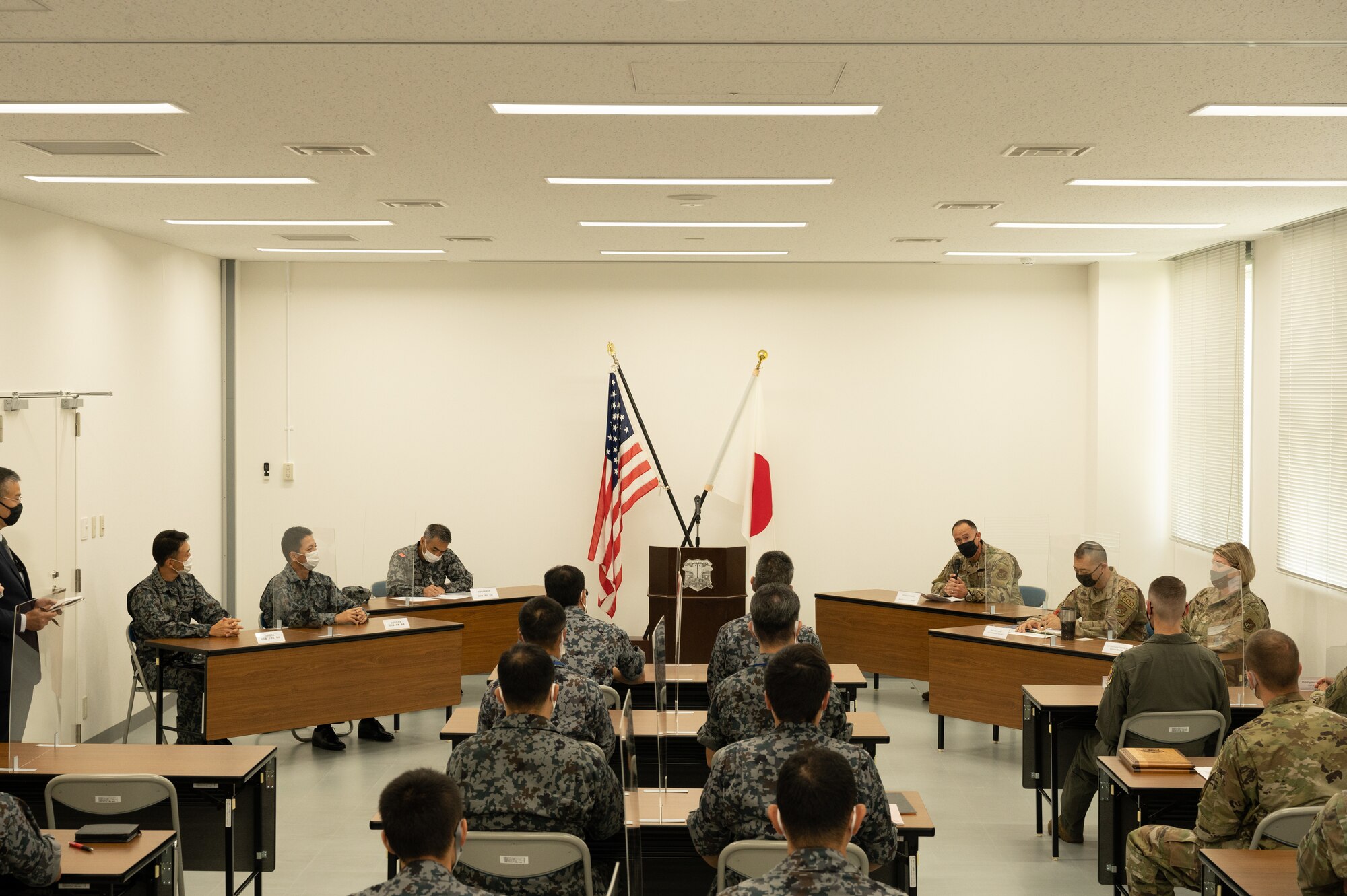 Several people in uniform are seated in a large room. At the front of the room are the American and Japanese flags.
