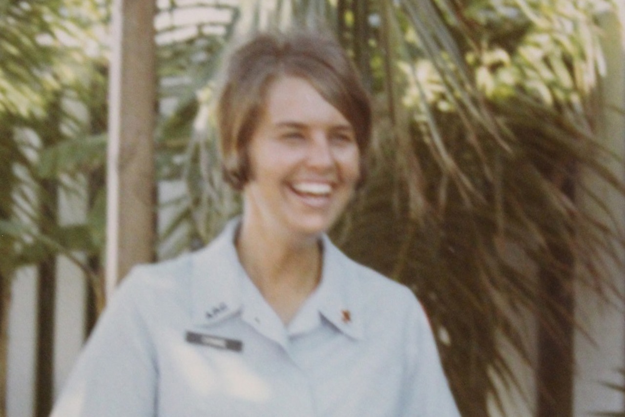 Woman smiles outside in front of tropical foliage.