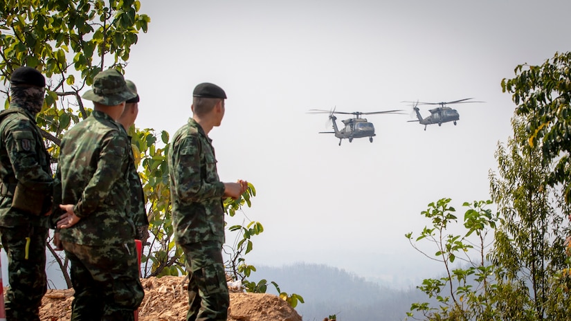 Military personnel on a mountain top, watch as two helicopters pass by.