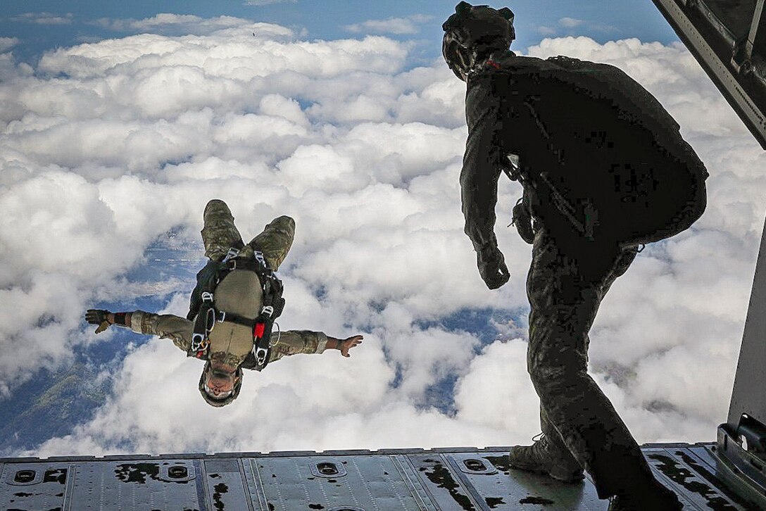 An airman freefalls upside down after jumping out of an airborne aircraft with clouds in the background.