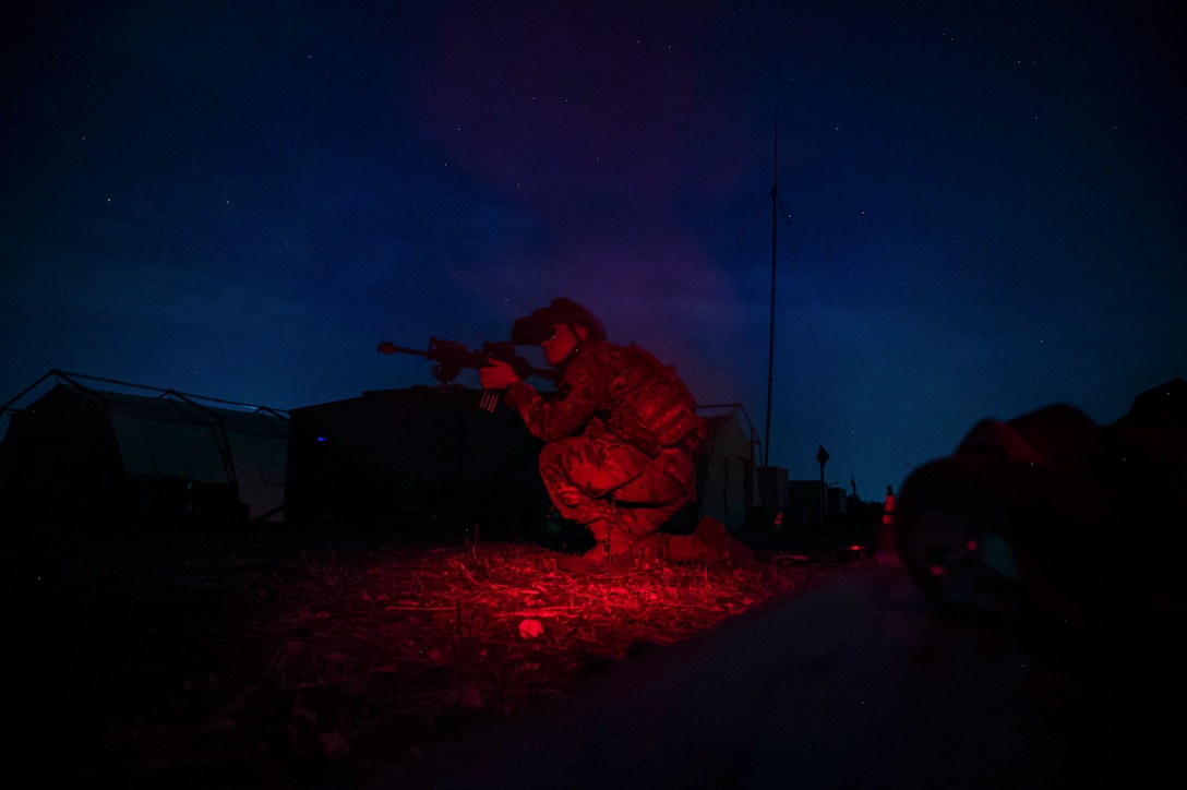 An airman aims a weapon in a field in the dark illuminated by a red light.