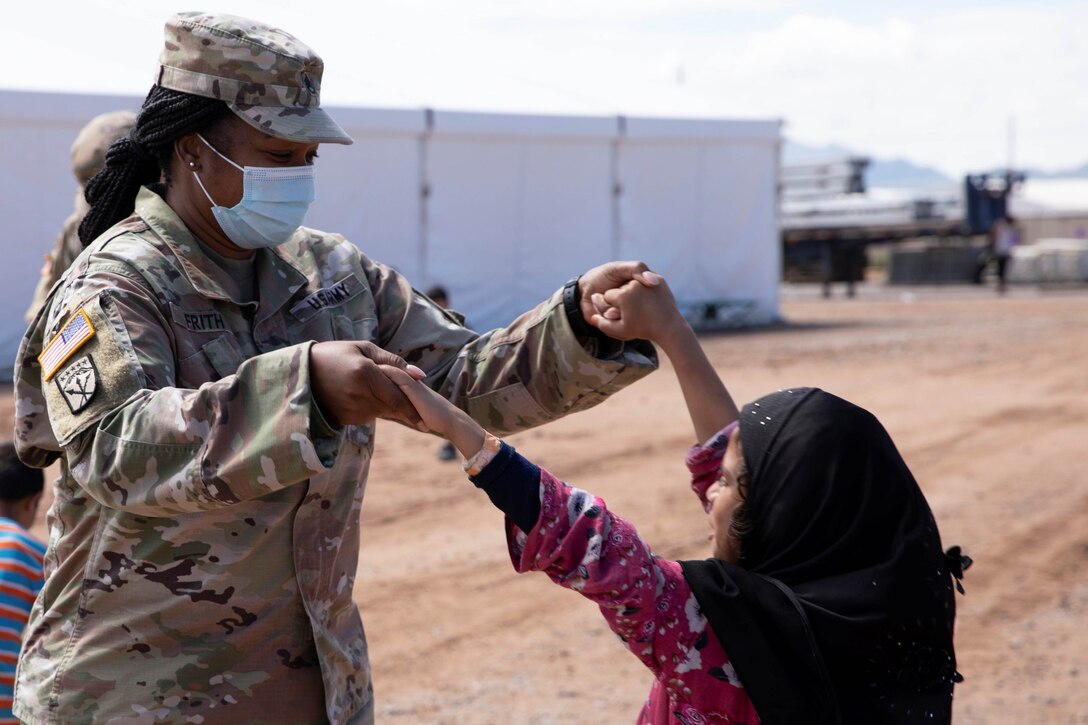 A female service member and a young girl in a head scarf hold hands.