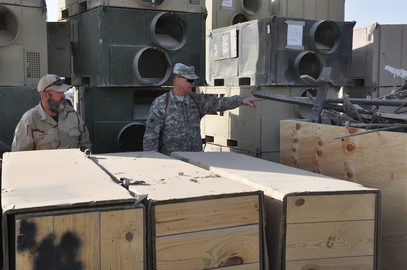Two men, one in uniform, stand amidst crates of supplies.