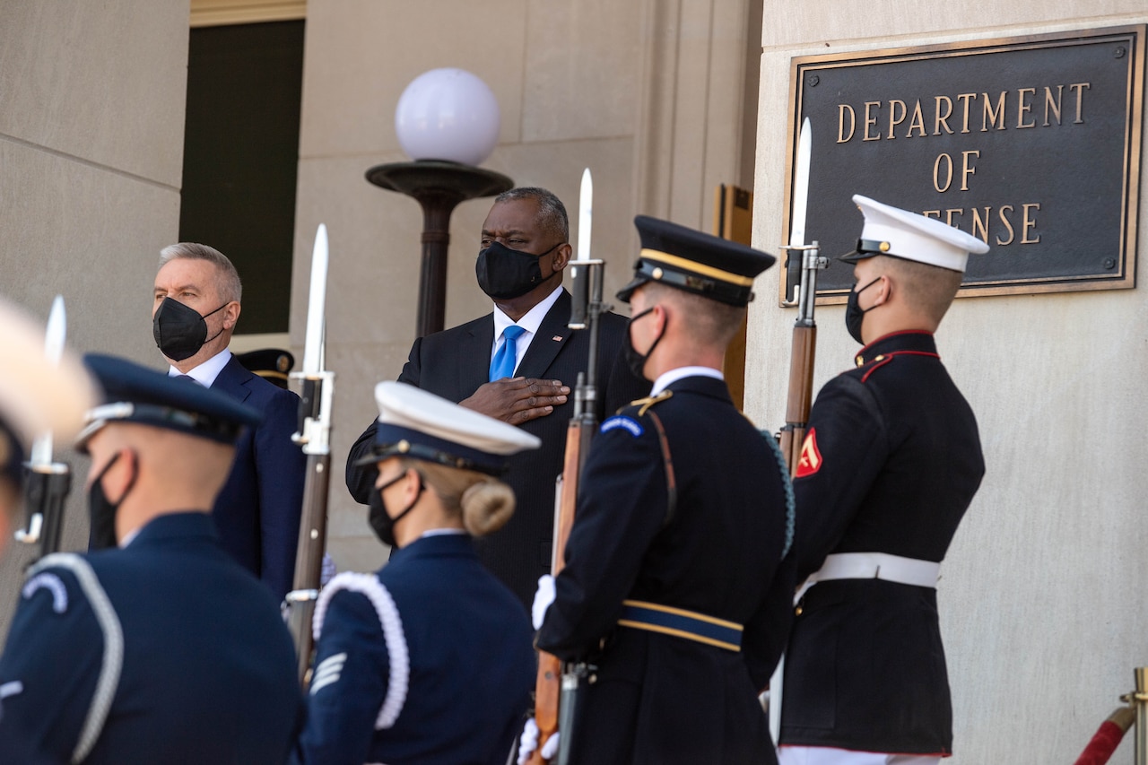 Defense secretary places hand over heart and stands with another official.