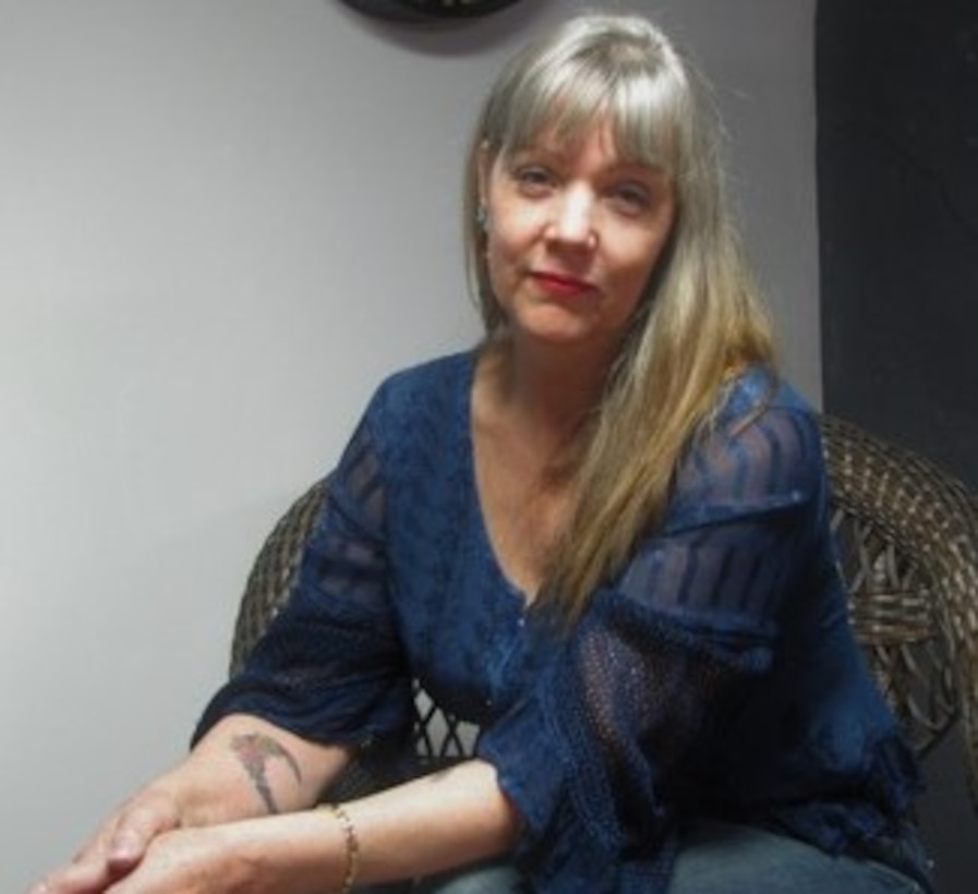 A woman poses with a calm face, wearing a blue dress, sitting in a wicker chair with hands folded toward her right.