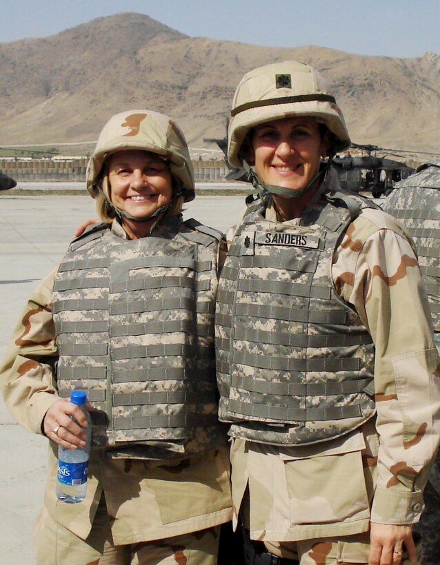 Two women wearing helmets, body armor and desert Army uniforms stand in front of mounts.