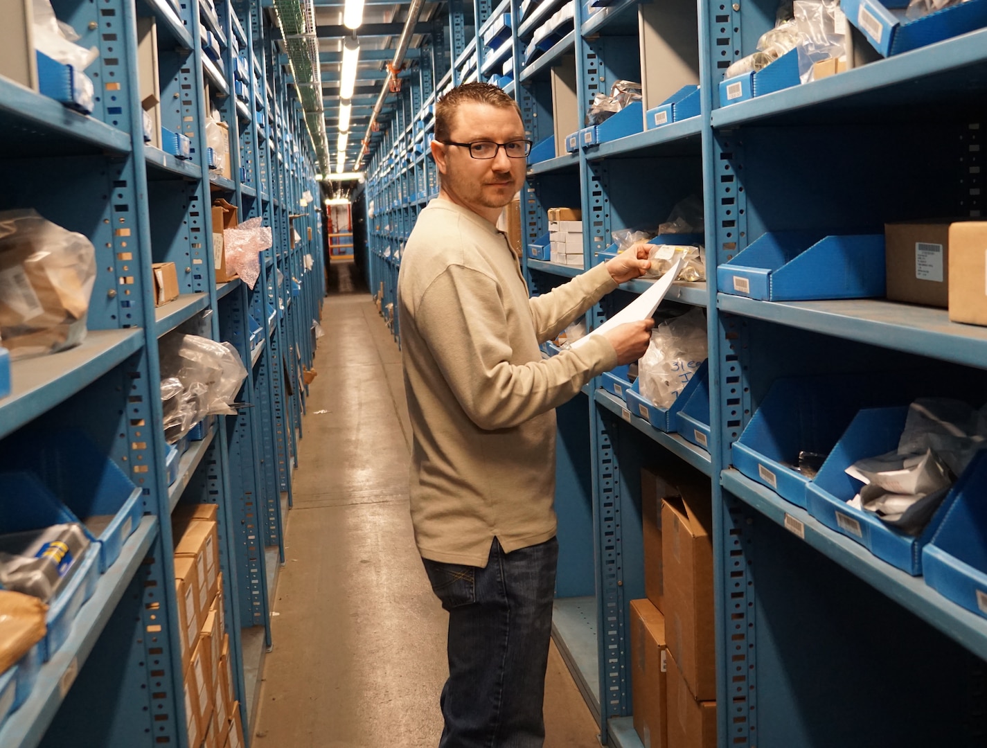 Guy in brown shirt and jeans stands in front of blue shelving with supplies holding a piece of paper in one hand.