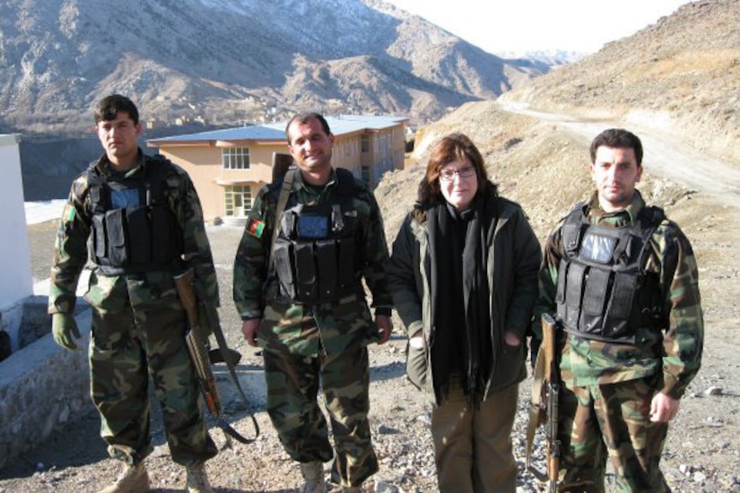 A woman and three men pose for a photo in rough terrain; mountains are in the background.