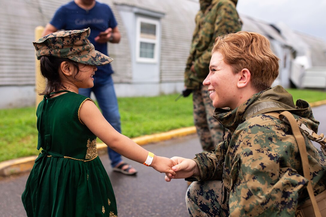 A Marine holds the hand of a smiling child wearing a Corps cover.