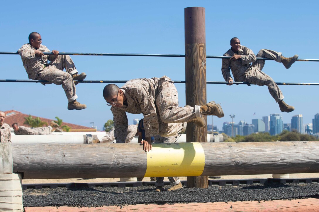 Two Marine Corps recruits pull themselves over bars as another jumps over a log.