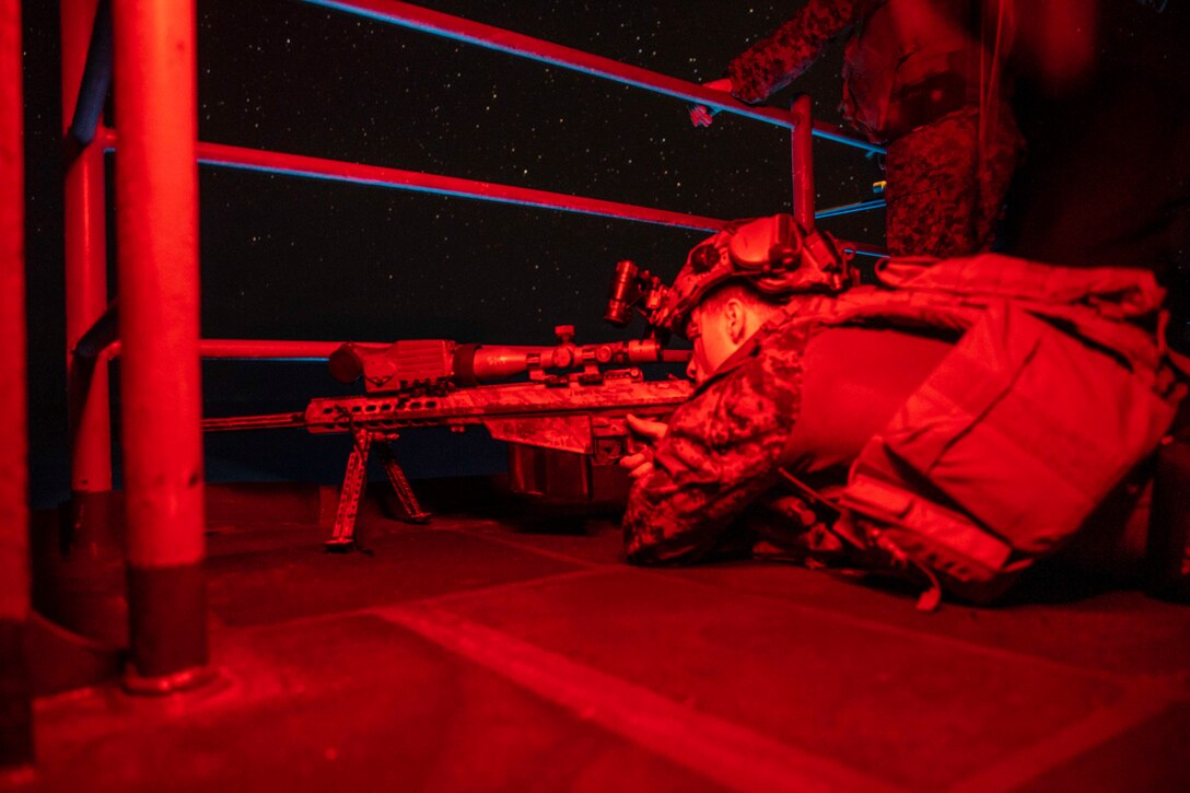 A Marine illuminated by red light lies on the deck of a ship looking through the scope of a weapon.