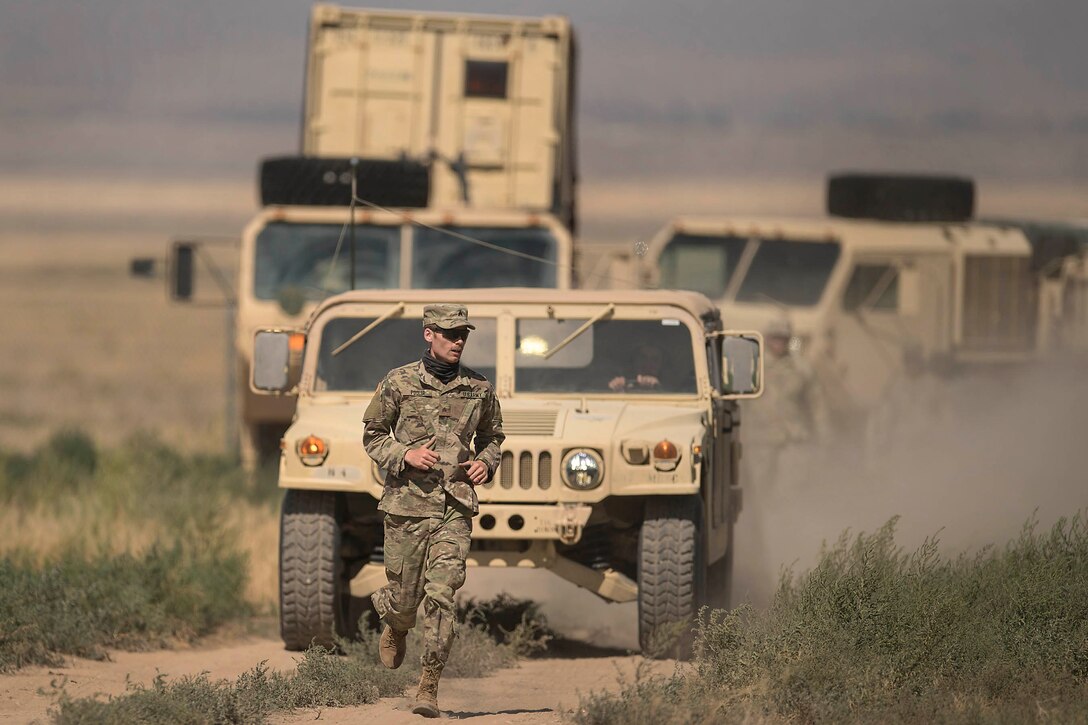 A soldier runs along a dirt road in front of military vehicles.