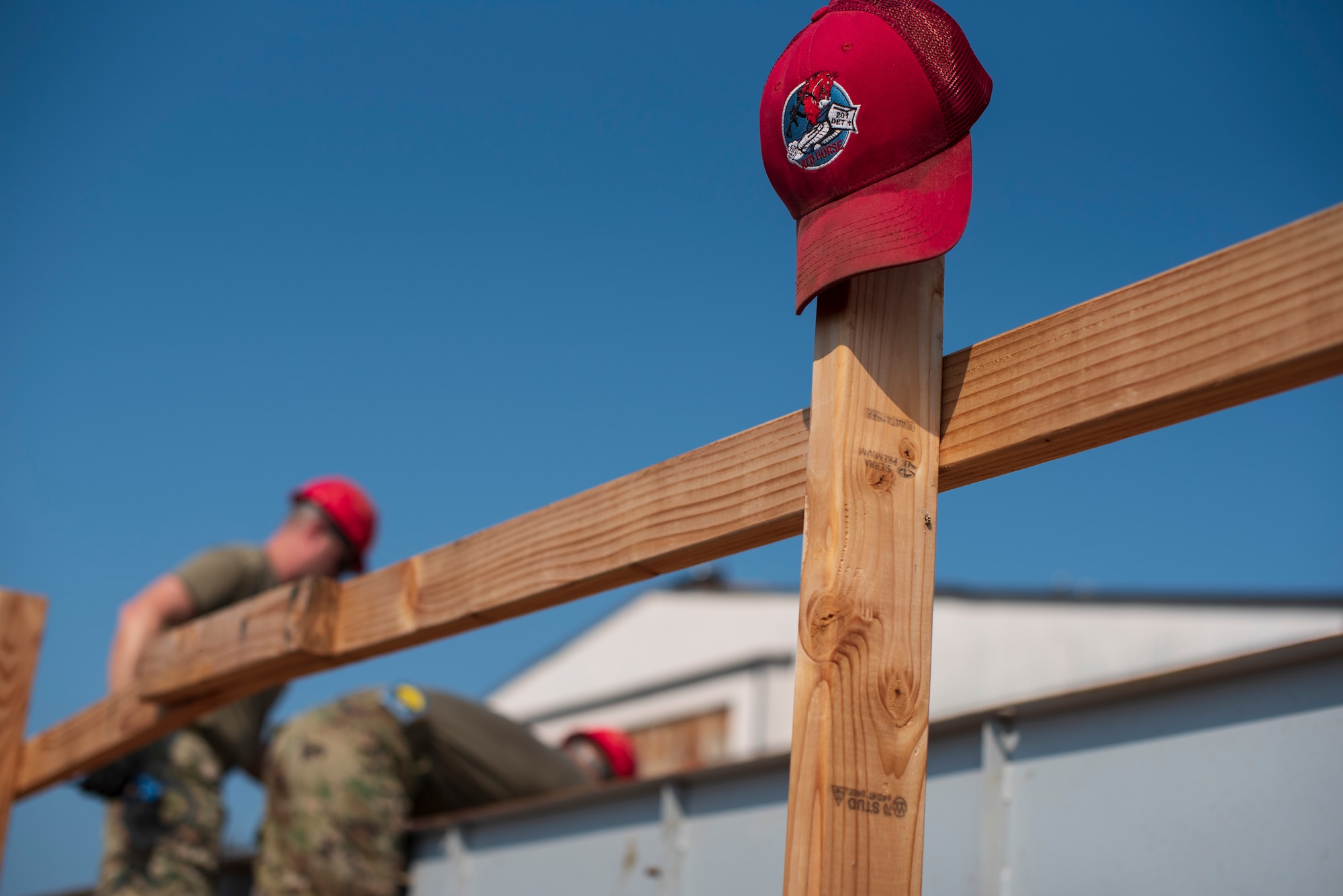 A red RED HORSE cap rests on a wooden rail while two men work in the background.