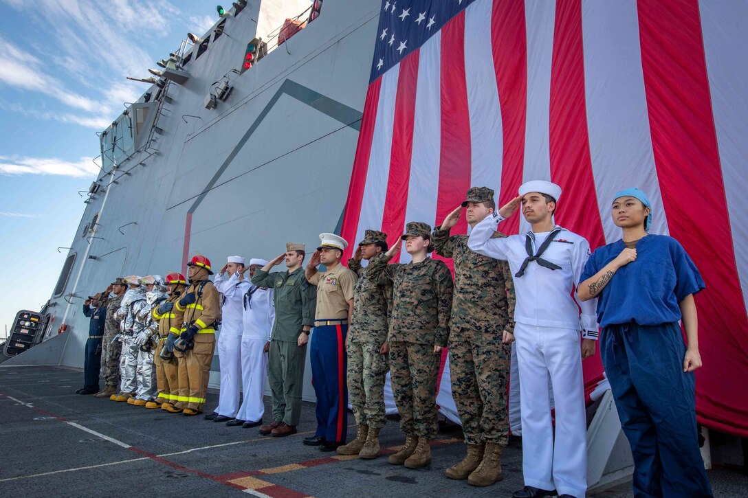 A line of service members salutes as they stand in front of a large U.S. flag.