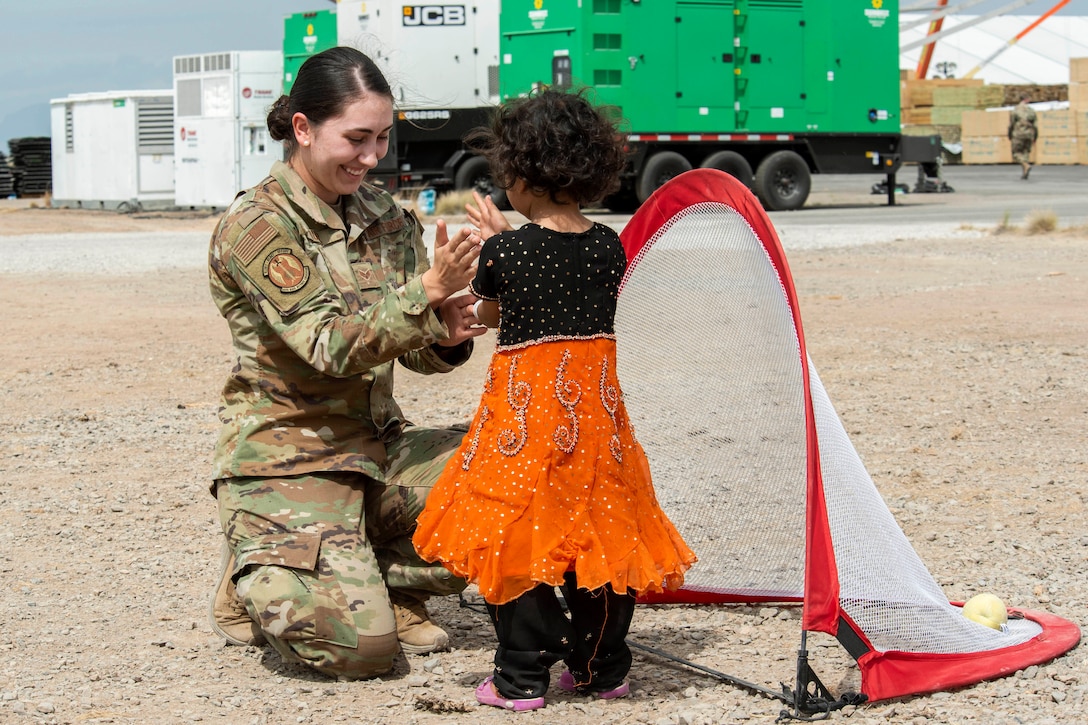 An airman plays a hand game with a child next to a goal post in a sandy area.