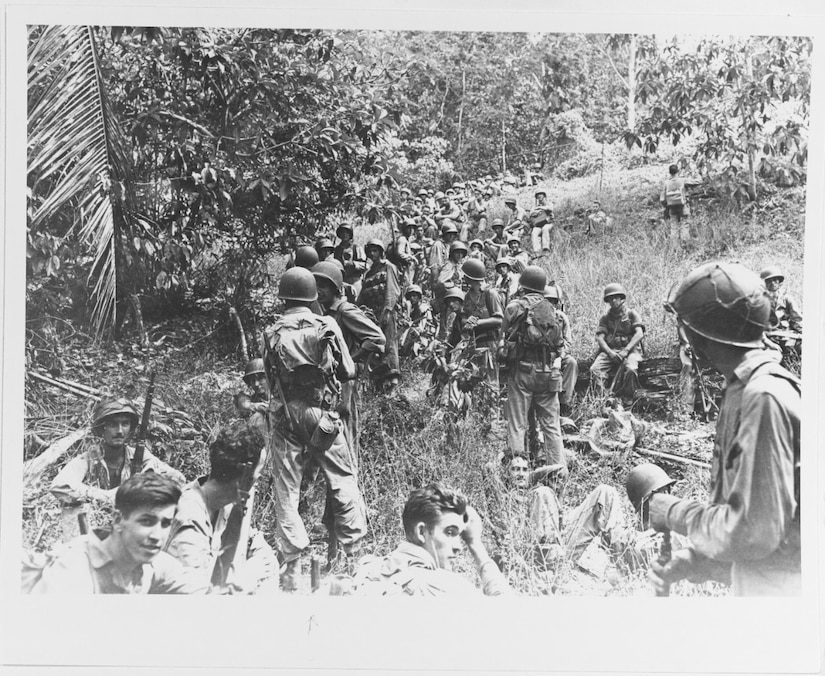 Dozens of men in combat gear walk along a jungle path. Some rest in the nearby brush.